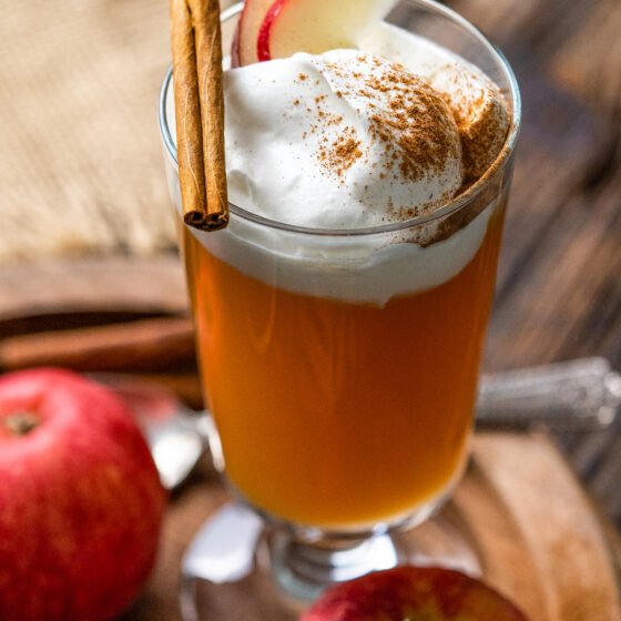 cocktail glass with apple cider and topped with whipped cream, apple slices, cinnamon, a piece of graham cracker and cinnamon stick