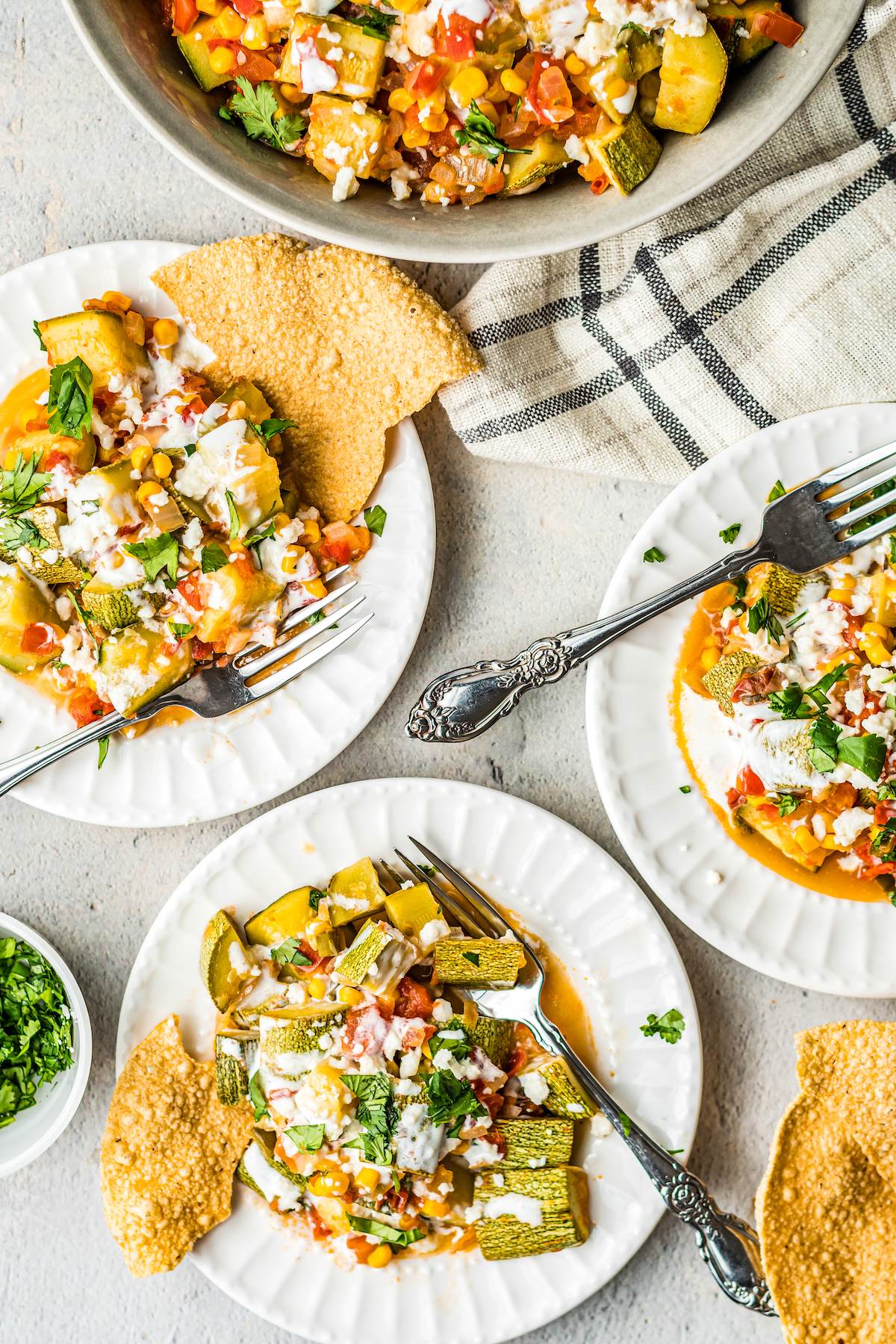 Overhead shot of a table with a large bowl of cooked summer squash with tomatoes and corn. Smaller servings have been scooped onto white plates.