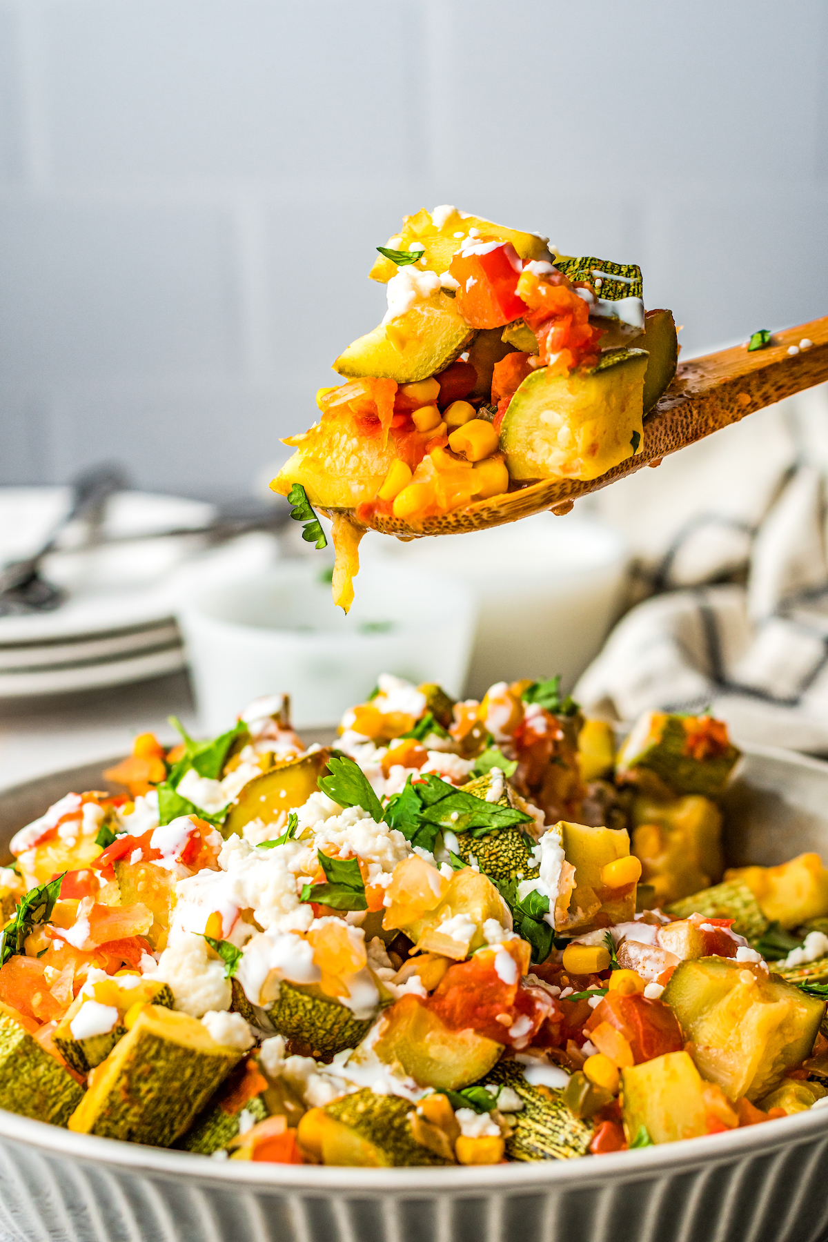 A wooden spoon scooping a serving of squash out of a large dish of veggies.