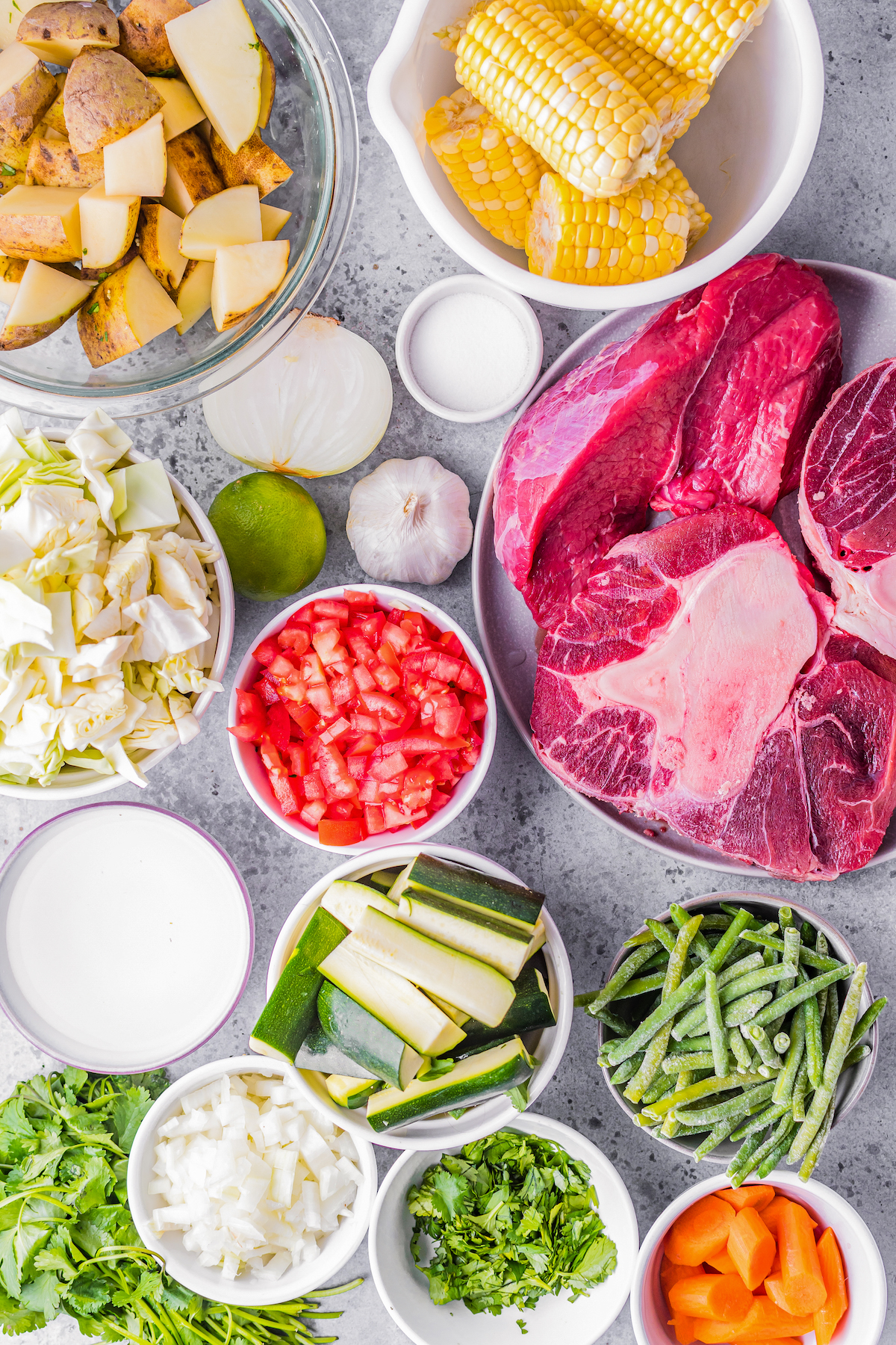 Caldo de Res ingredients being prepped including beef, tomatoes, carrots, green beans, zucchini, herbs, onions, cabbage, lime, and garlic