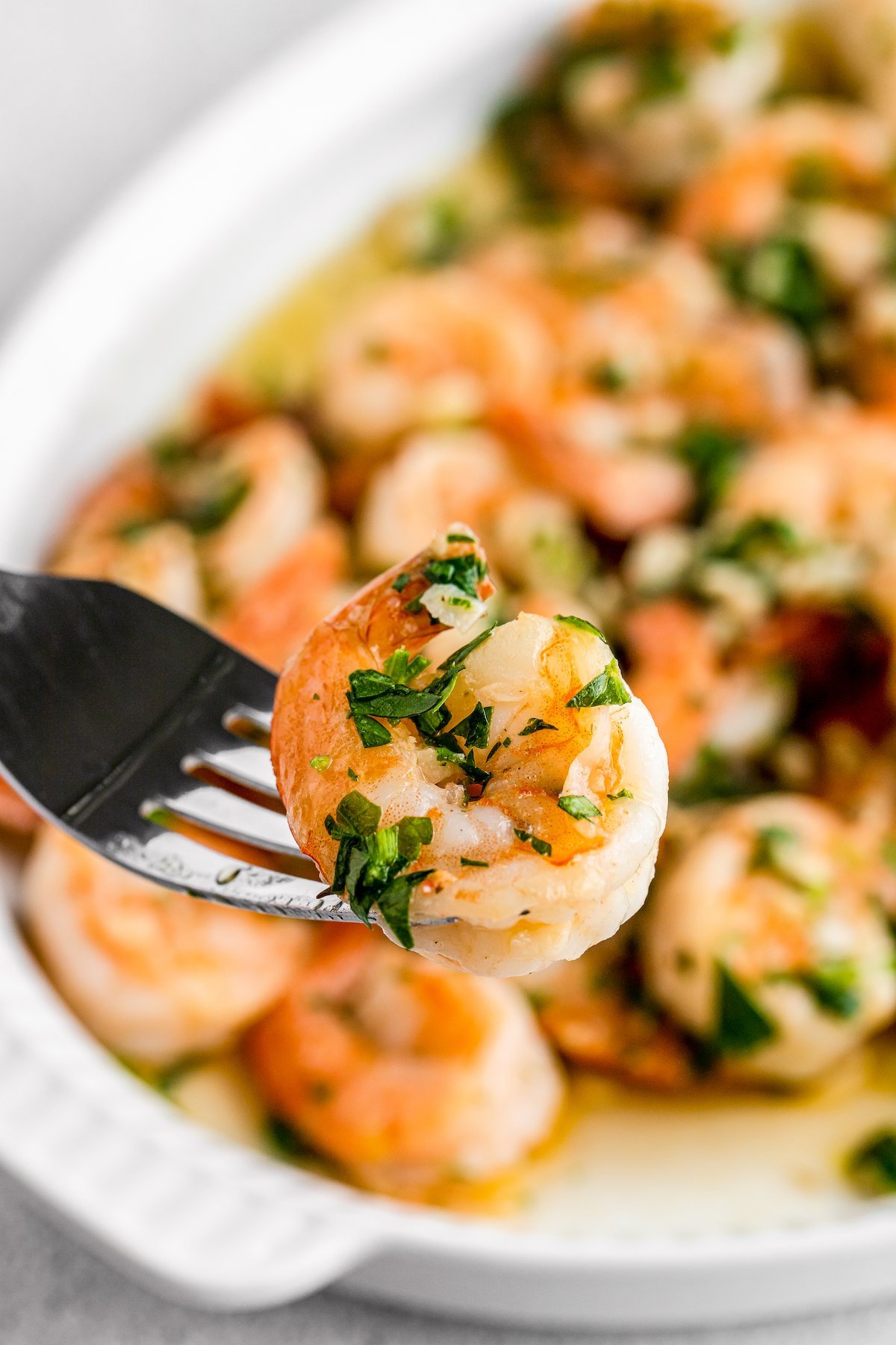 A single sauteed shrimp on a fork, held toward the camera. A plate of garlic shrimp is in the background of the shot.