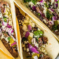 Three tacos stuffed with carne asada and topped with red onion, cheese crumbles and cilantro.