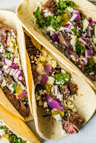 Three tacos stuffed with carne asada and topped with red onion, cheese crumbles and cilantro.