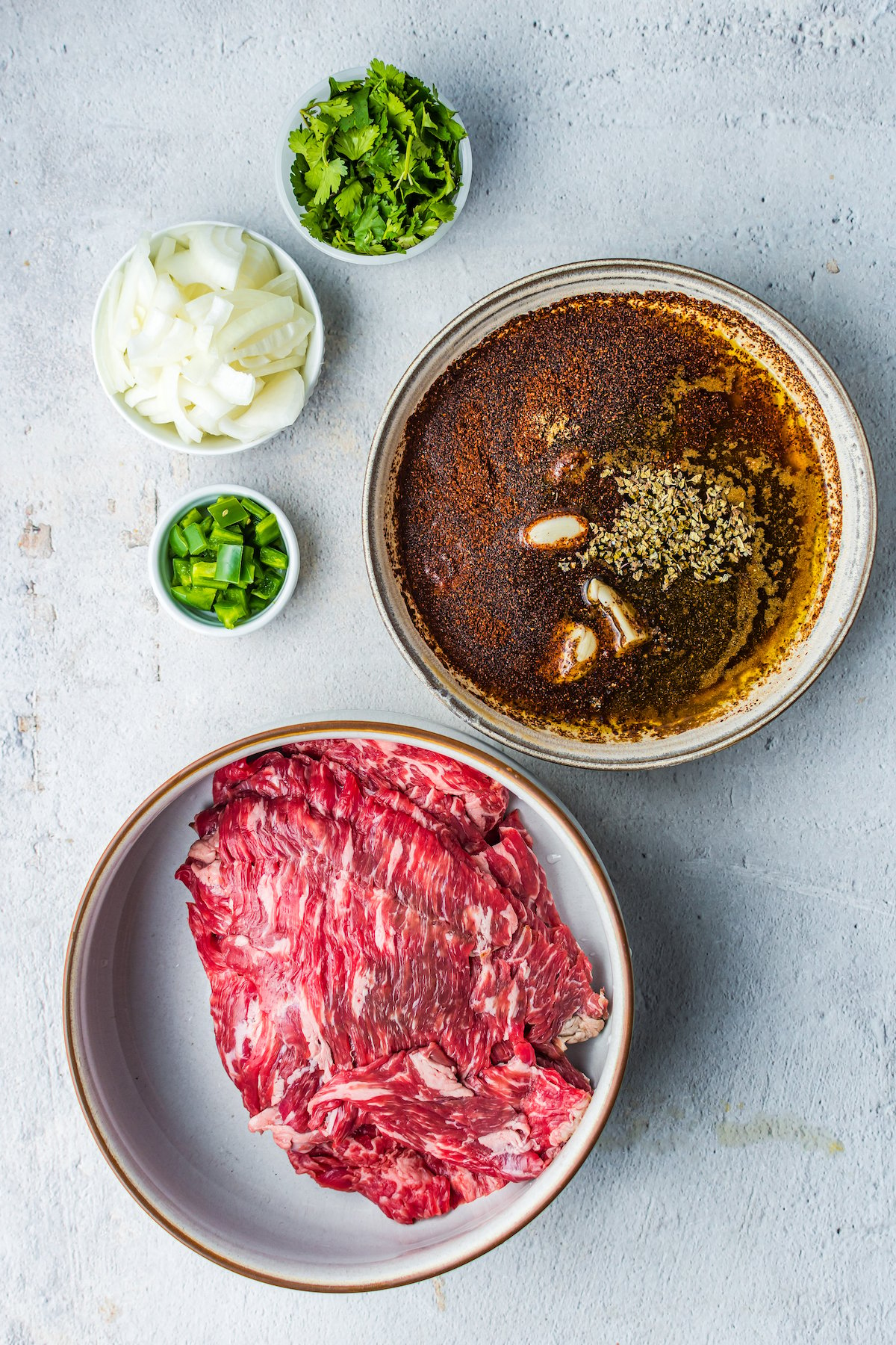 A dish of flank steak next to a bowl of marinade ingredients, a dish of jalapeno, a dish of chopped onion, and a dish of cilantro.