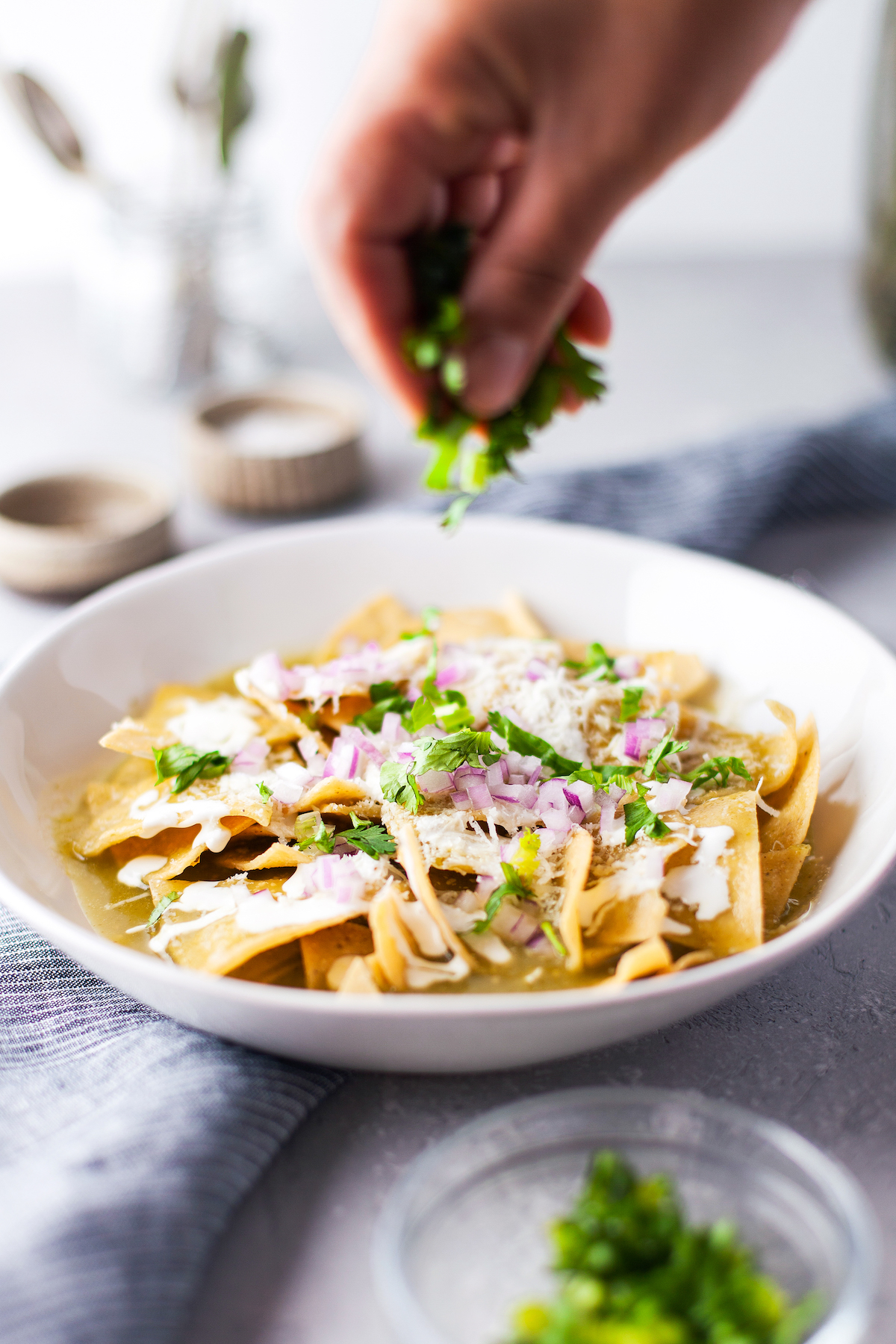 tortilla chips with salsa verde on top being garnished with cilantro, red onions, and cheese in a white bowl