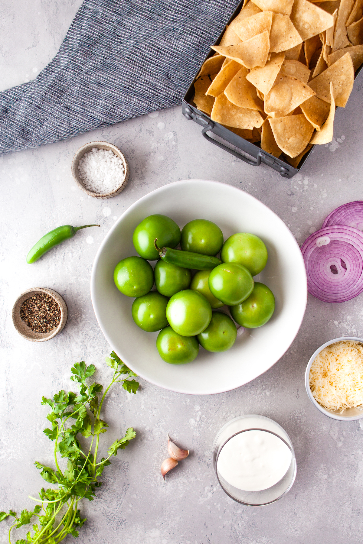 tomatillos in a bowl with other salsa verde ingredients like salt, red onions, pepper, and cilantro on the counter next to it