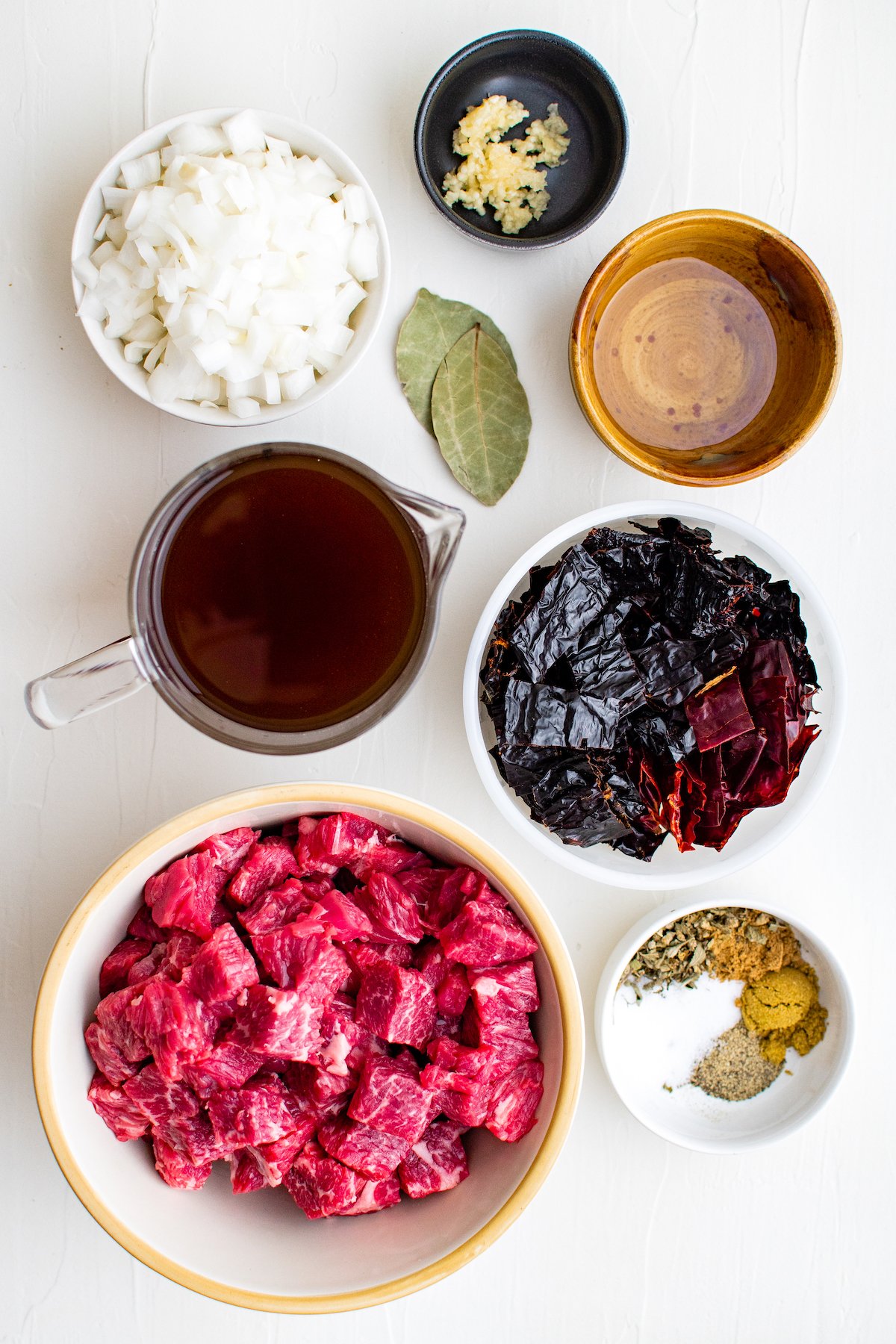 Ingredients in prep bowls to make Chili Colorado like dried peppers, beef broth, diced onions, garlic, spices, and cut up beef pieces 