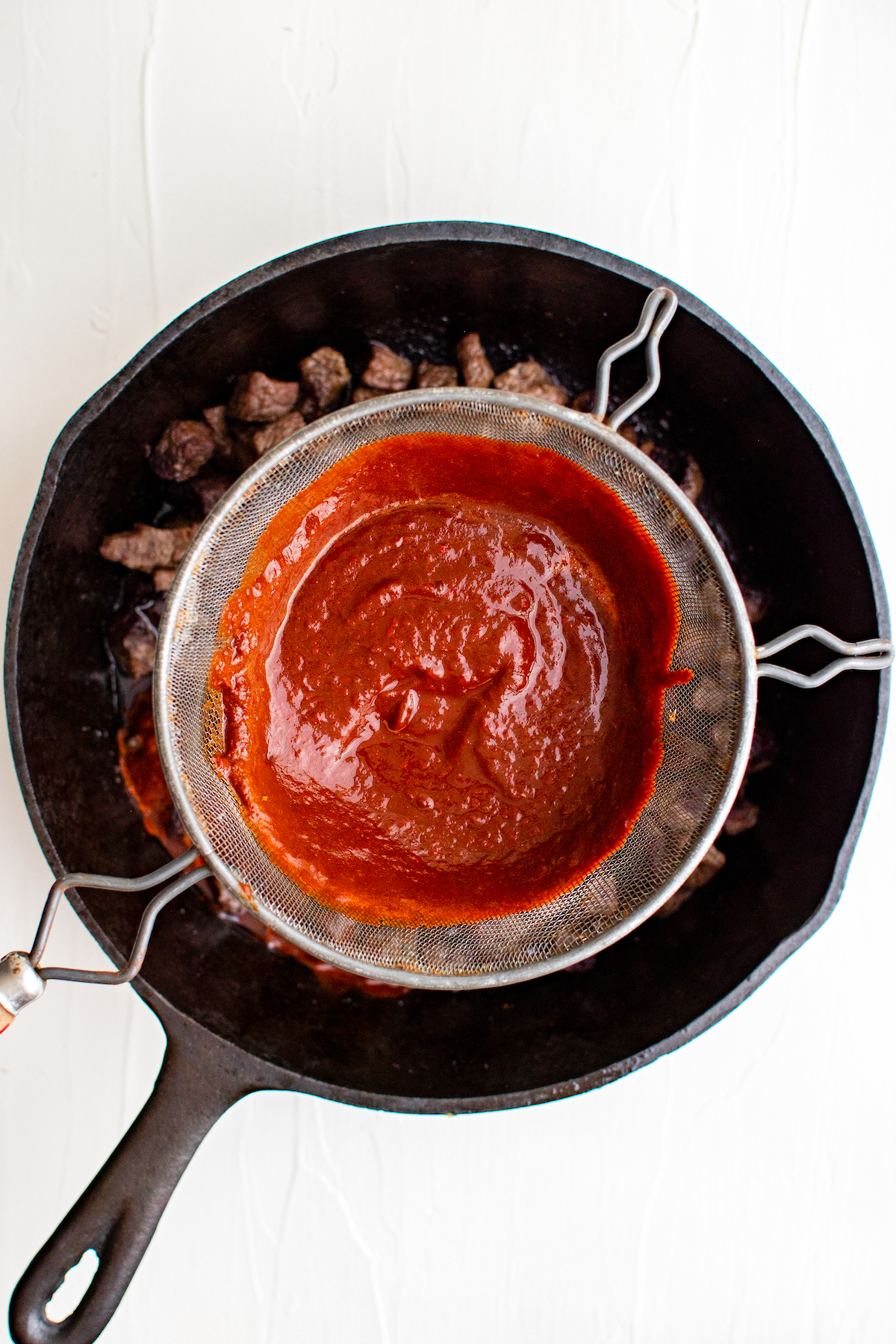 Red chili-based sauce being strained added to Chili Colorado in a cast iron skillet 