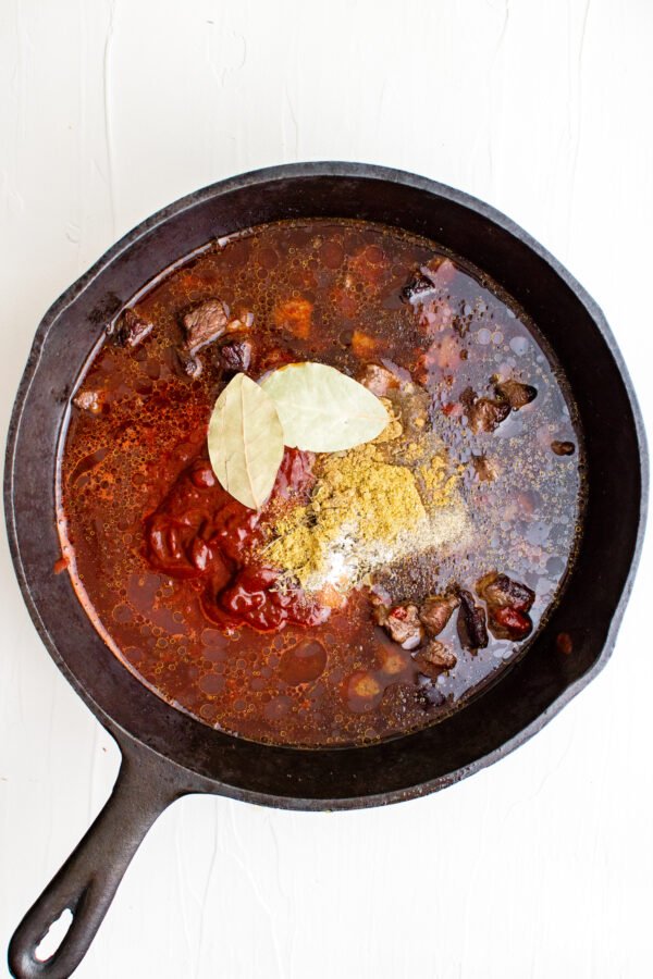 A cast iron skillet with the ingredients for Mexican beef chili with red sauce including spices and beef