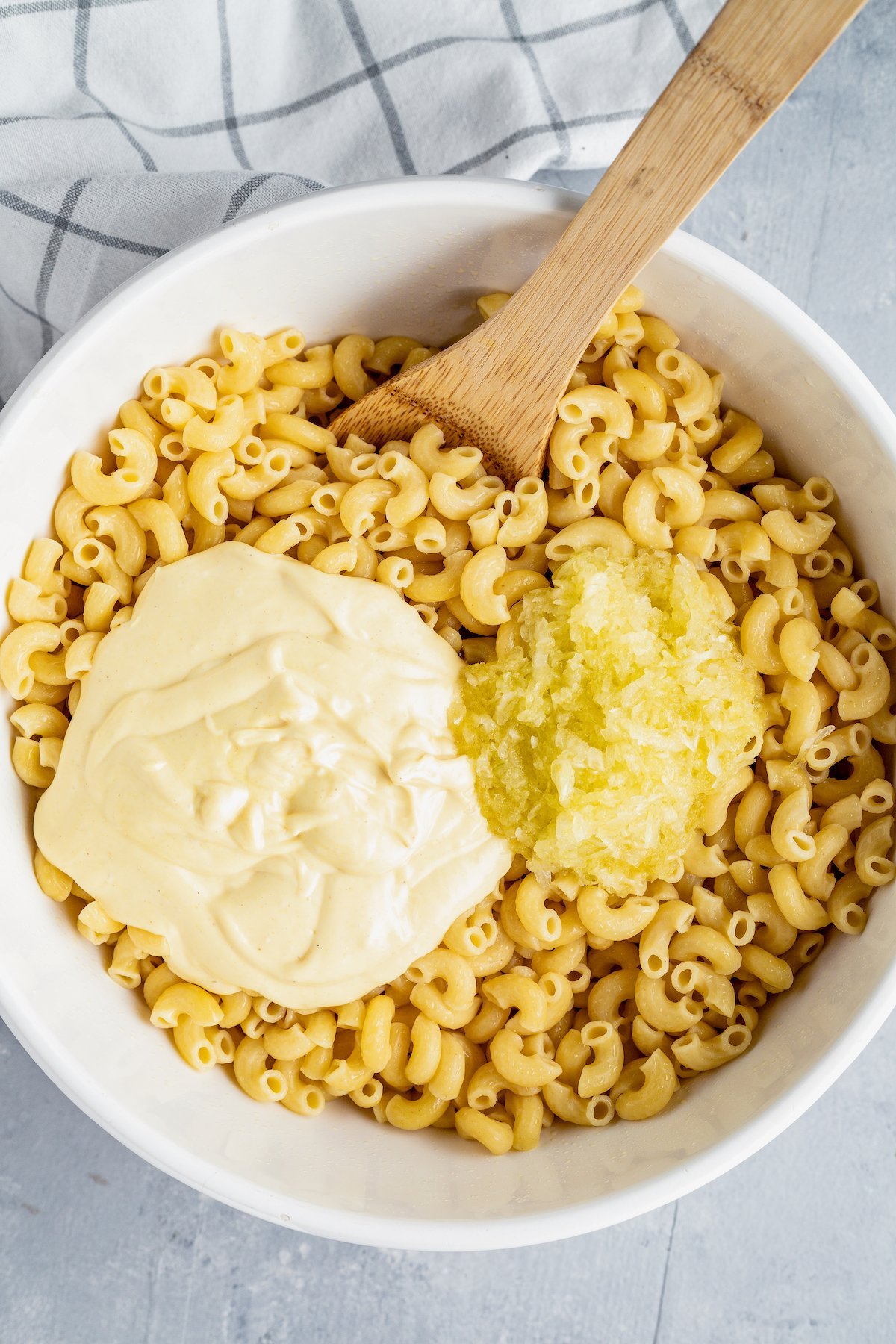 Creamy dressing and grated onion being stirred into a bowl of cooked elbow pasta.