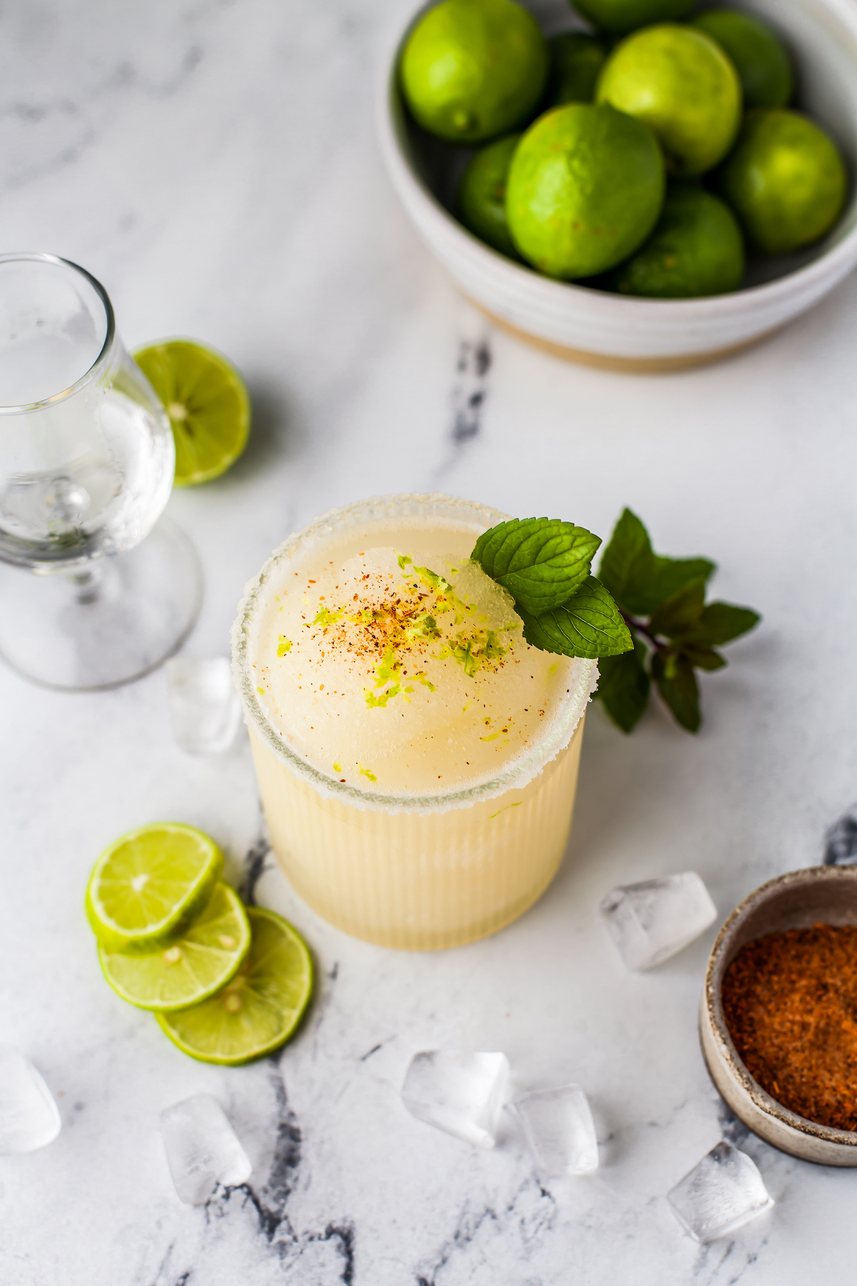 a frozen margarita in a glass with mint and chili spice garnish on a table with lime slices, ice cubes, and a small bowl of seasonings
