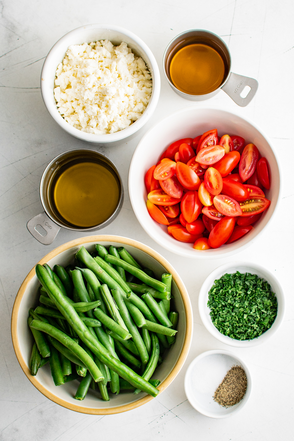 ingredients in prep bowls to make green bean salad, like green beans, grape tomatoes, feta cheese, olive oil, and parsley