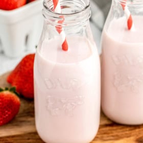 A glass of milk with strawberry syrup on a cutting board with strawberries.