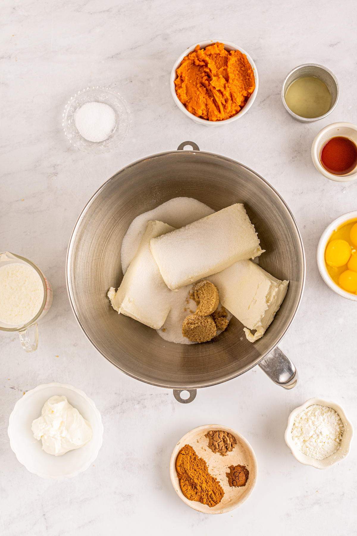 cream cheese, sugar, and brown sugar in a mixing bowl surrounded by prep bowls filled with various ingredients to make pumpkin cheesecake