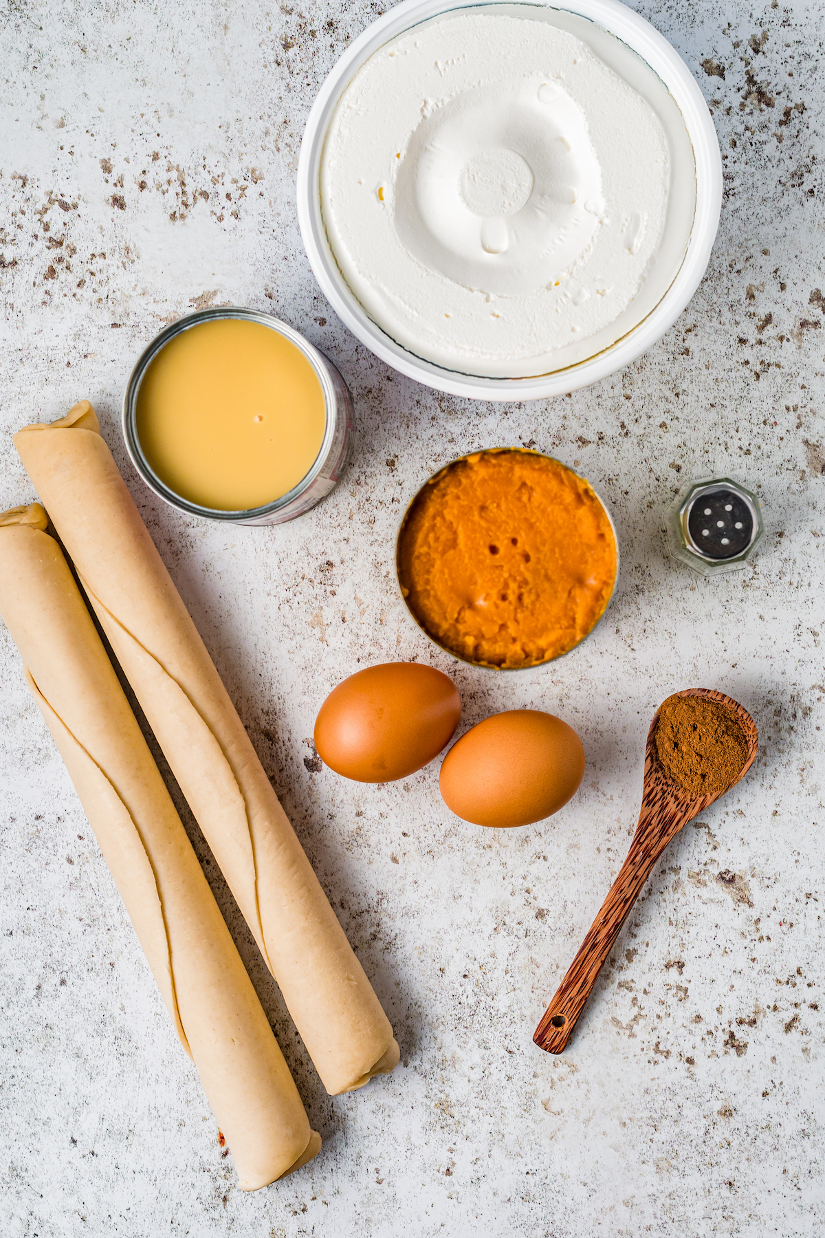 ingredients to make pumpkin pie on a countertop incluiding rolled pastry dough, pumpkin puree, eggs, pumpkin pie spice, sweetened condensed milk, and cool whip