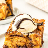 A slice of croissant bread pudding with nutella on a white plate with ice cream and chocolate sauce on top.