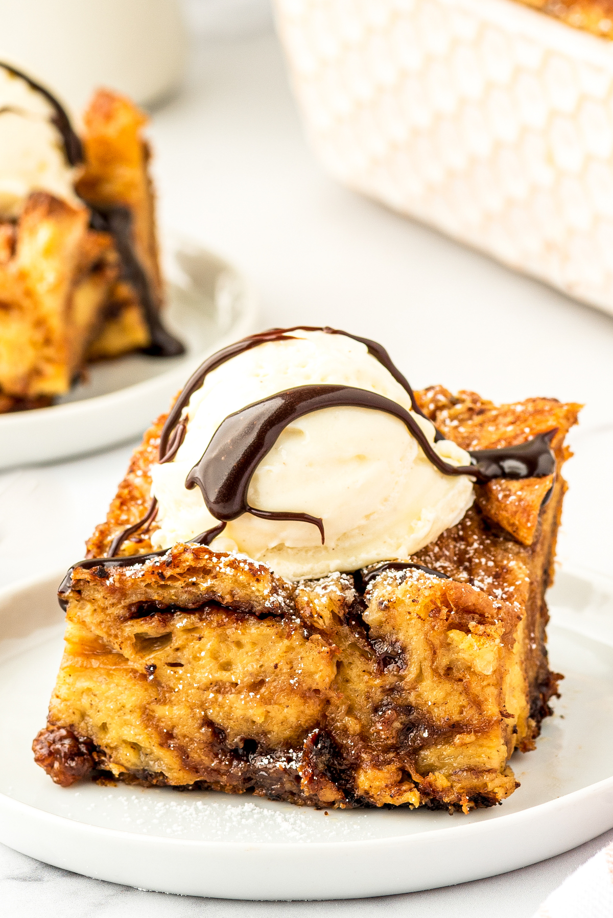 A slice of croissant bread pudding with nutella on a white plate with ice cream and chocolate sauce on top.