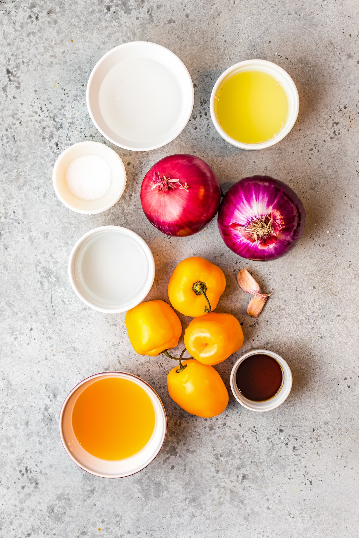 Red onions, habanero peppers, and garlic next to prep bowls containing salt, vinegar, maple syrup, apple cider vinegar, and lime juice