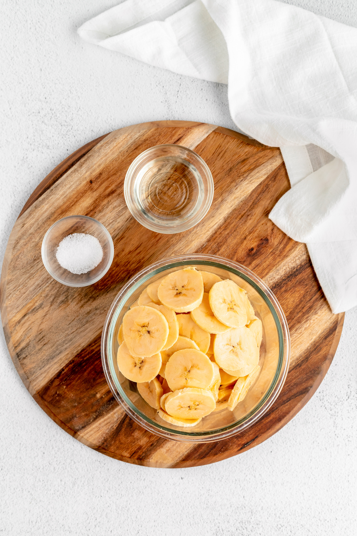 Overhead shot of a wooden cutting board. A glass bowl of sliced plantains, and two small glass dishes of salt and oil, are arranged on the cutting board.