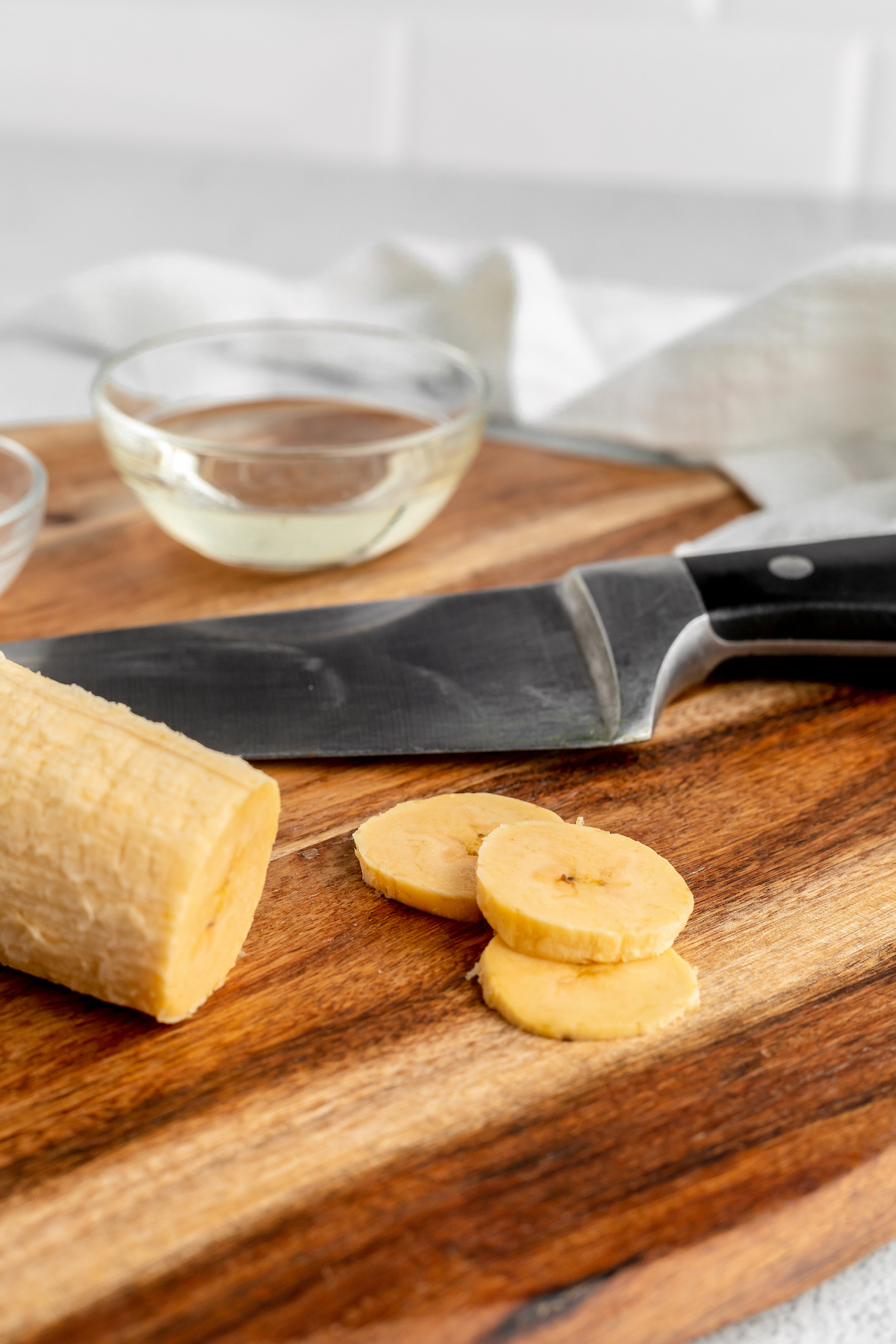 A peeled plantain on a cutting board with a sharp knife. Several slices have been cut from the plantain and are lying next to the knife.