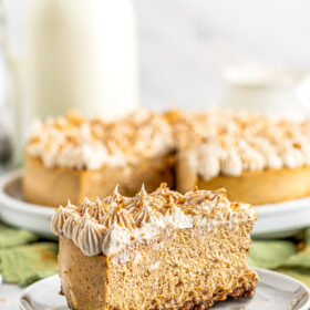 A stack of small dessert plates, with one slice of pumpkin cheesecake with gingersnap crust on the top plate. In the background is a whole cheesecake with one slice cut out, and a bottle of milk.