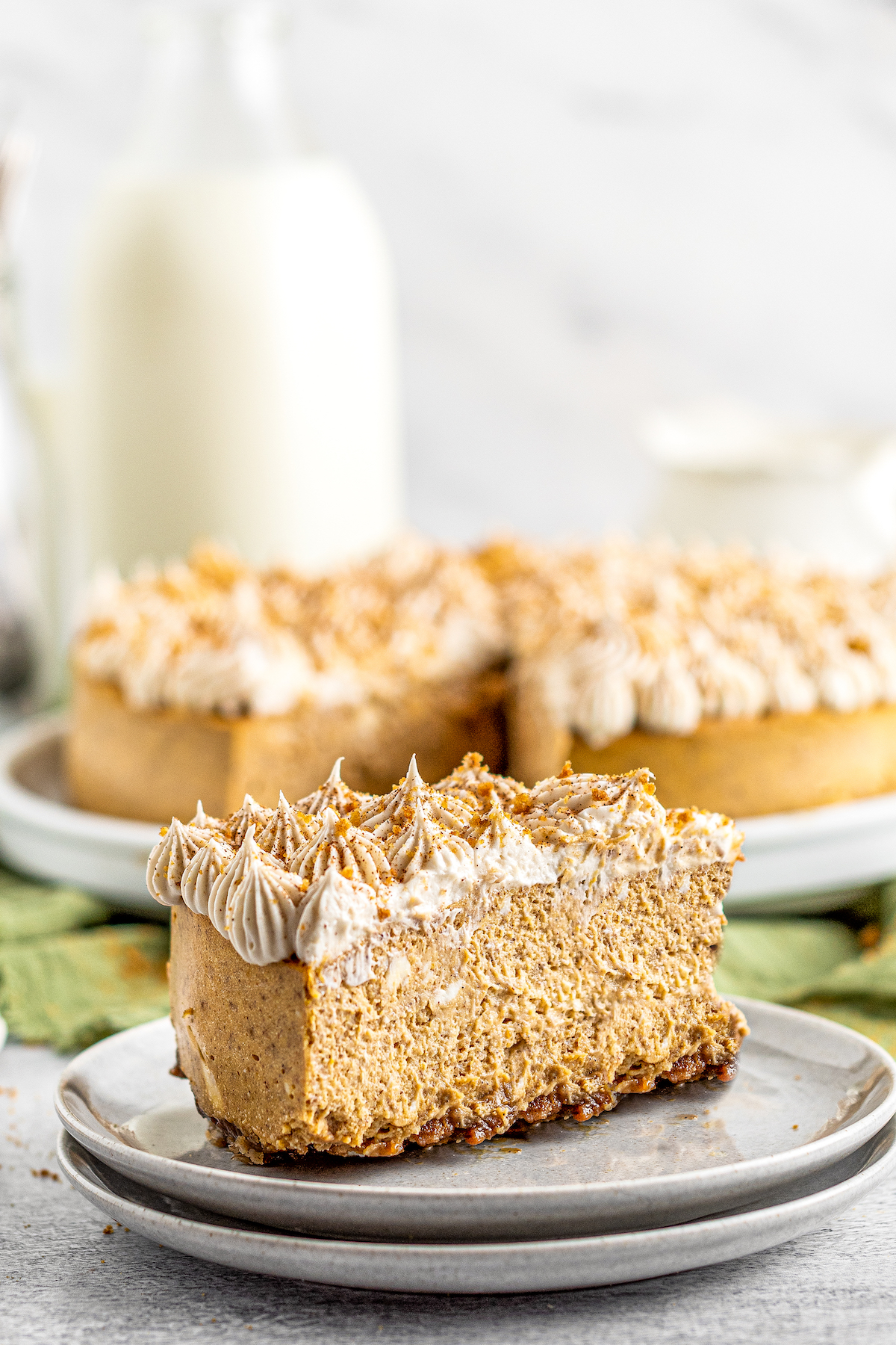 A stack of small dessert plates, with one slice of pumpkin cheesecake with gingersnap crust on the top plate. In the background is a whole cheesecake with one slice cut out, and a bottle of milk.