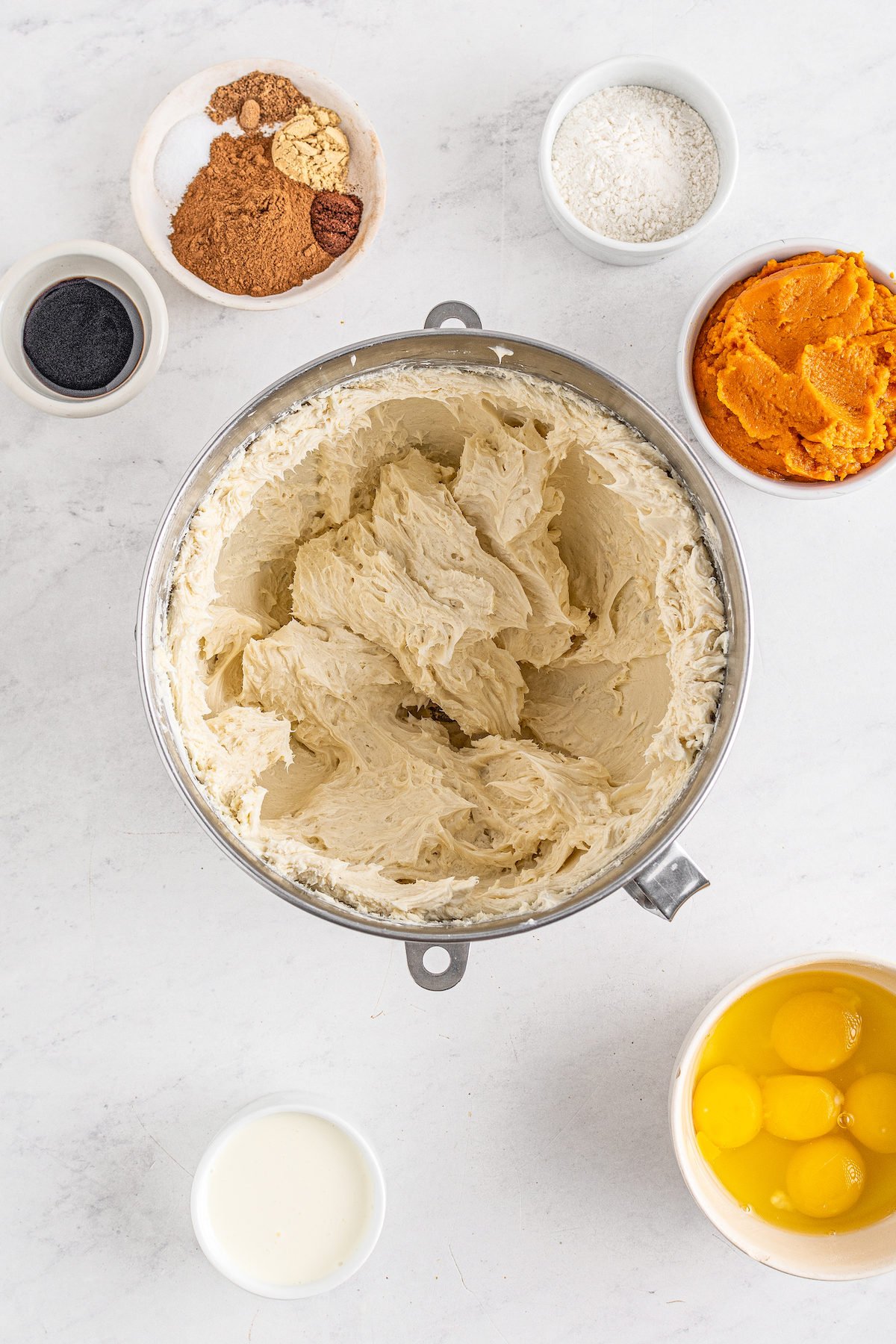 A mixing bowl with beaten cream cheese inside. Smaller dishes of raw eggs, pumpkin puree, vanilla, spices, and sugar are arranged around the mixing bowl.