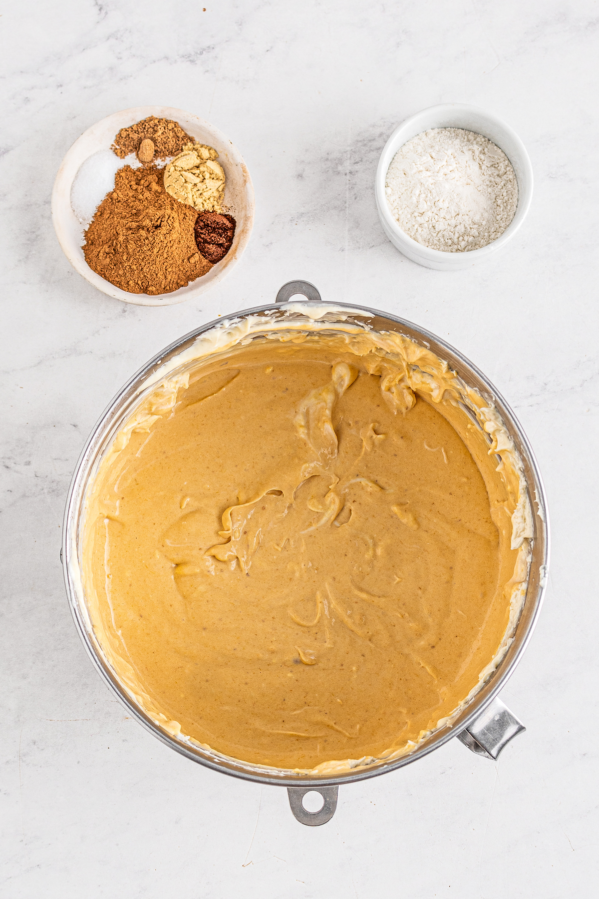 A large mixing bowl with creamy, pumpkin batter inside. Smaller dishes of ingredients are sitting nearby.