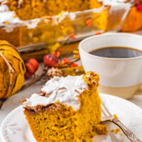 A serving of pumpkin cinnamon roll cake with vanilla glaze, next to a cup of black coffee. In the background of the shot are autumn decorations and a glass baking dish with the rest of the cake.