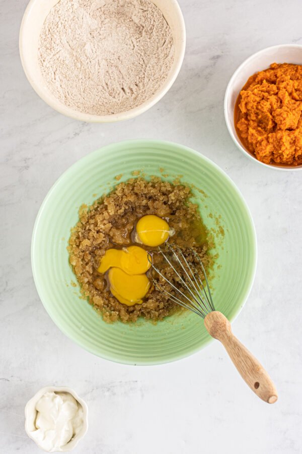 Brown sugar and eggs in a mixing bowl. A dish of pumpkin puree and a dish of dry ingredients are nearby on the work surface.
