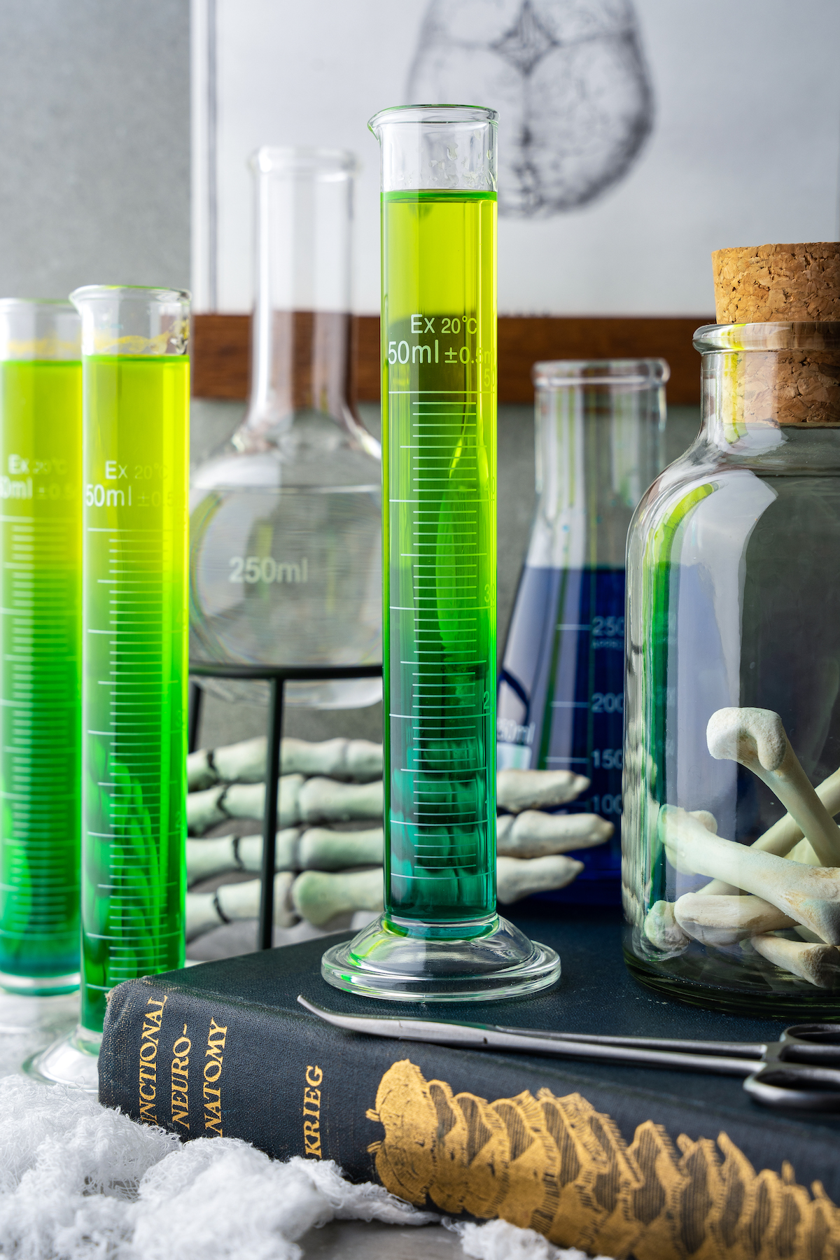 three shots of liquor that gradually turn from green to yellow in tall graduated cylinders on a table with Halloween decorations