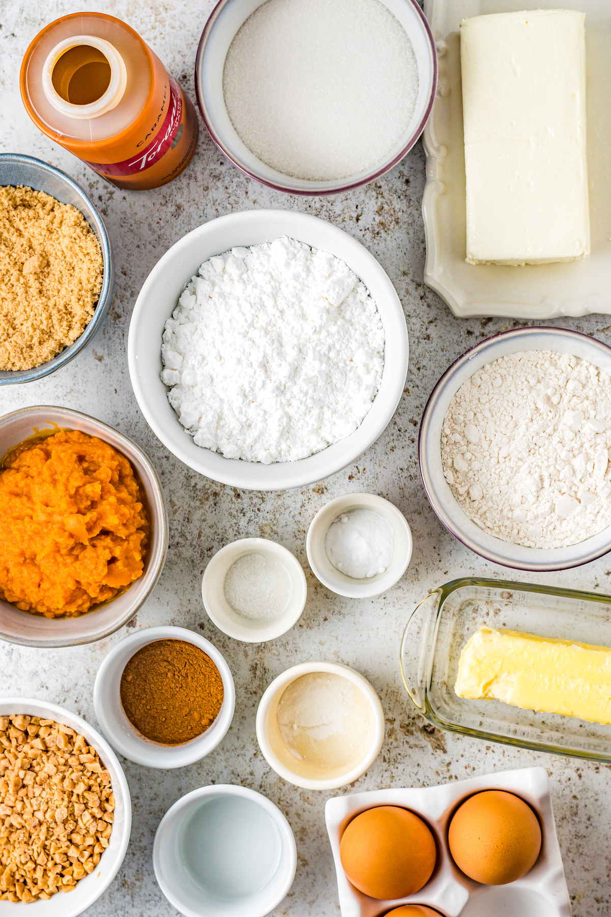 From top left: Caramel sauce, white sugar, cream cheese, brown sugar, powdered sugar, flour, pumpkin, baking soda, baking powder, butter, toffee bits, vanilla, salt, pumpkin pie spice, eggs.