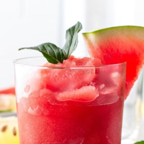 A glass of watermelon frosé with a watermelon wedge and fresh basil on top.