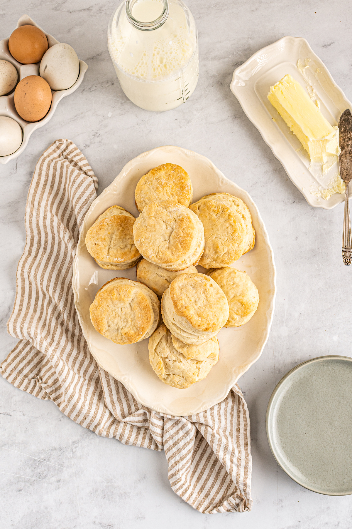 An oval platter of homemade biscuits next to a plate of butter, a carton of eggs, and a glass bottle of milk.