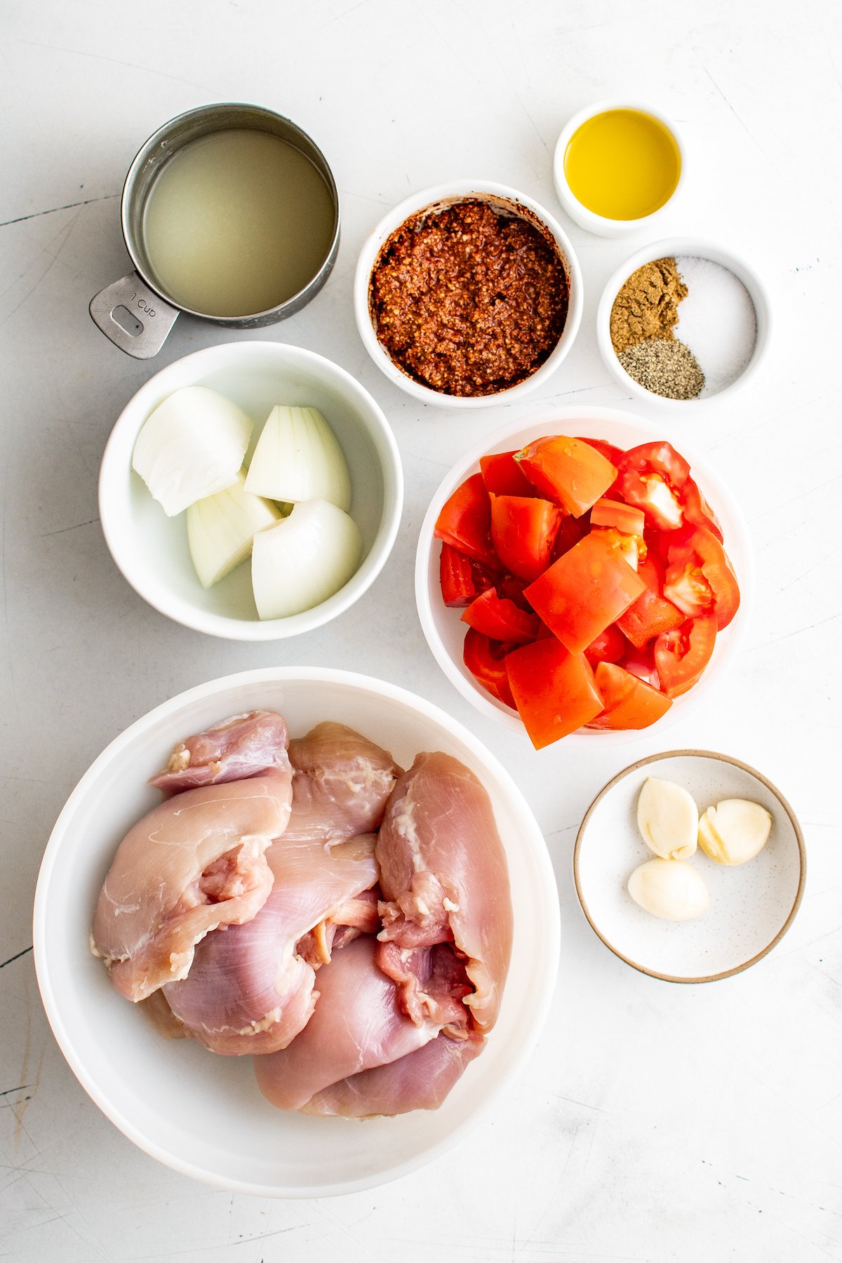 A bowl of raw chicken, a bowl of tomatoes, a bowl of garlic, a bowl of onion, a bowl of achiote paste, a bowl of seasonings, a bowl of oil, and a cup of lime juice