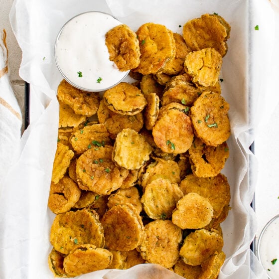 A paper-lined serving dish piled with fried pickles. A cup of ranch is also in the dish, with one pickle partially dipped in the ranch.