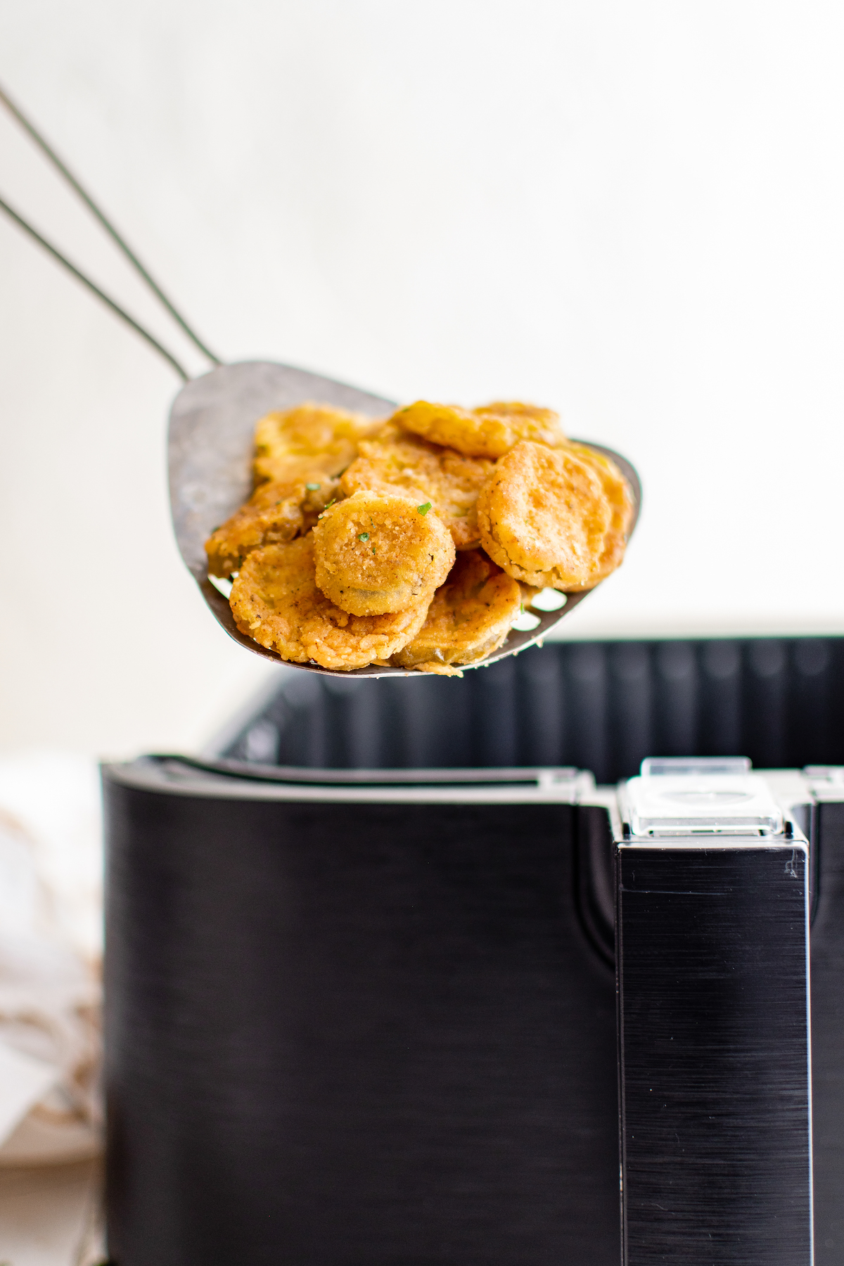 A metal slotted spatula lifting fried pickles out of an air fryer.