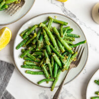 Air fryer green beans on a dinner plate with a fork. Lemon wedges and other plates of green beans are arranged artistically on the table.
