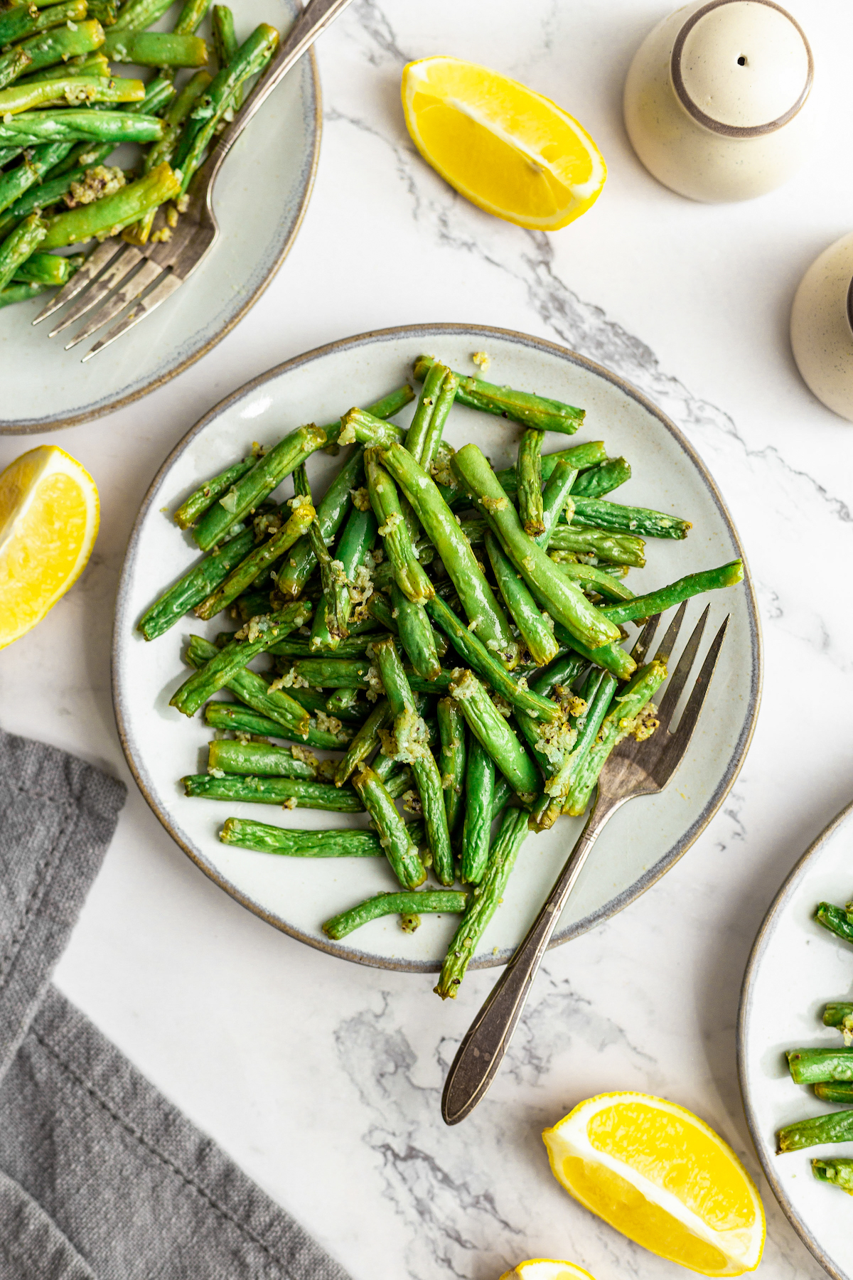 Air fryer green beans on a dinner plate with a fork. Lemon wedges and other plates of green beans are arranged artistically on the table.