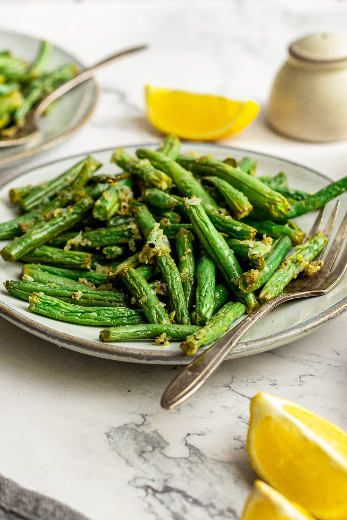 Cooked green beans with garlic. Lemon wedges and other plates of green beans are visible in the shot, along with salt and pepper shakers.