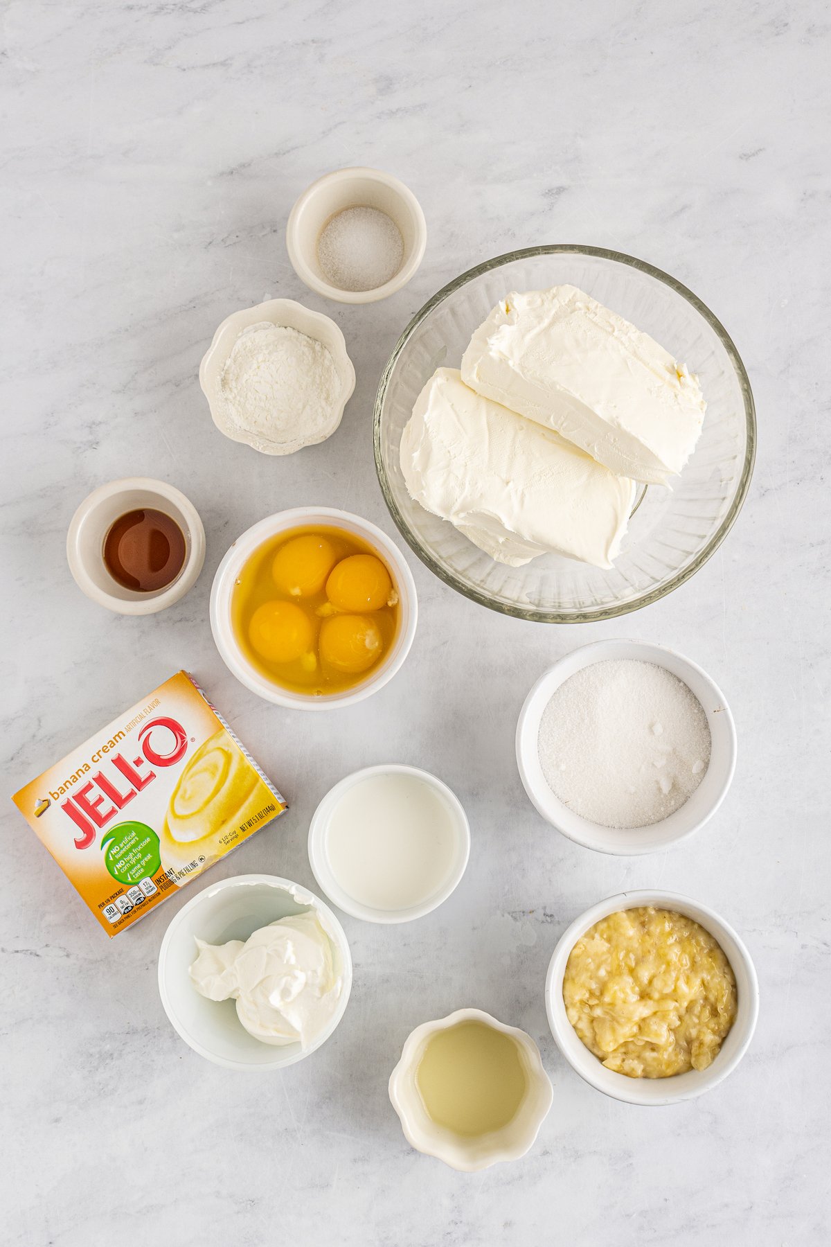 A bowl of cream cheese, a bowl of eggs, a bowl of mashed bananas, a bowl of heavy cream, a bowl of granulated sugar, a bowl of sour cream, a bowl of lemon juice, a bowl of vanilla extract, a bowl of salt, a bowl of cornstarch, and a Jell-O packed of banana cream pudding