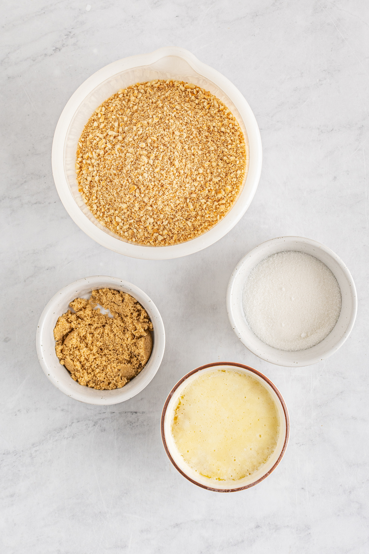 A bowl of vanilla wafer crumbs, a bowl of sugar, a bowl of brown sugar, and a bowl of melted butter