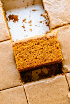 A square of pumpkin spice cake, turned sideways in the cake pan. The remaining pieces are still in their places, frosting-side-up.