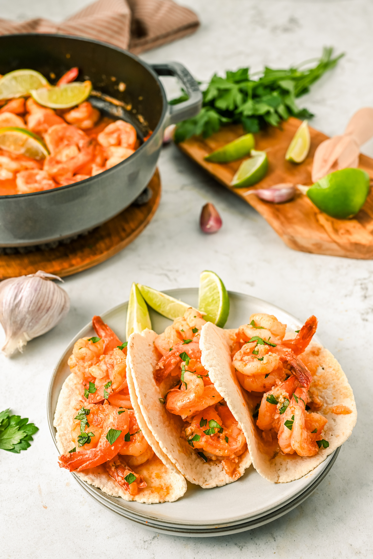 A plate of shrimp tacos with lime slices, on a table next to a head of garlic, lime slices, and a bowl of Camarones al Mojo de Ajo