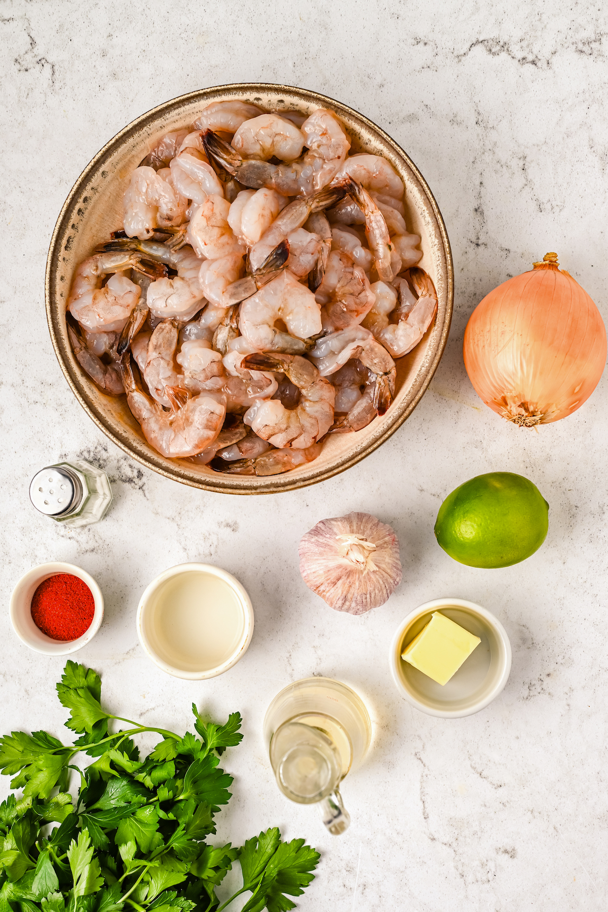 A bowl of raw shrimp, an onion, a lime, a head of garlic, a bowl of butter, a bowl of oil, a jar of vinegar, a bowl of paprika, a salt shaker, and a sprig of parsley