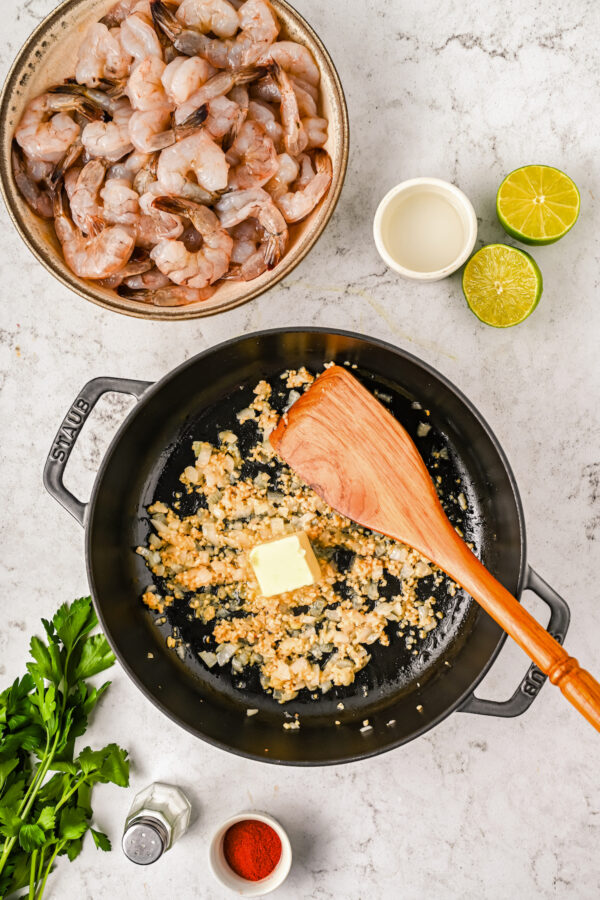 Onion and garlic cooking in a pot with a knob of unmelted butter and a wooden spoon, with raw shrimp, lime, and paprika on the side