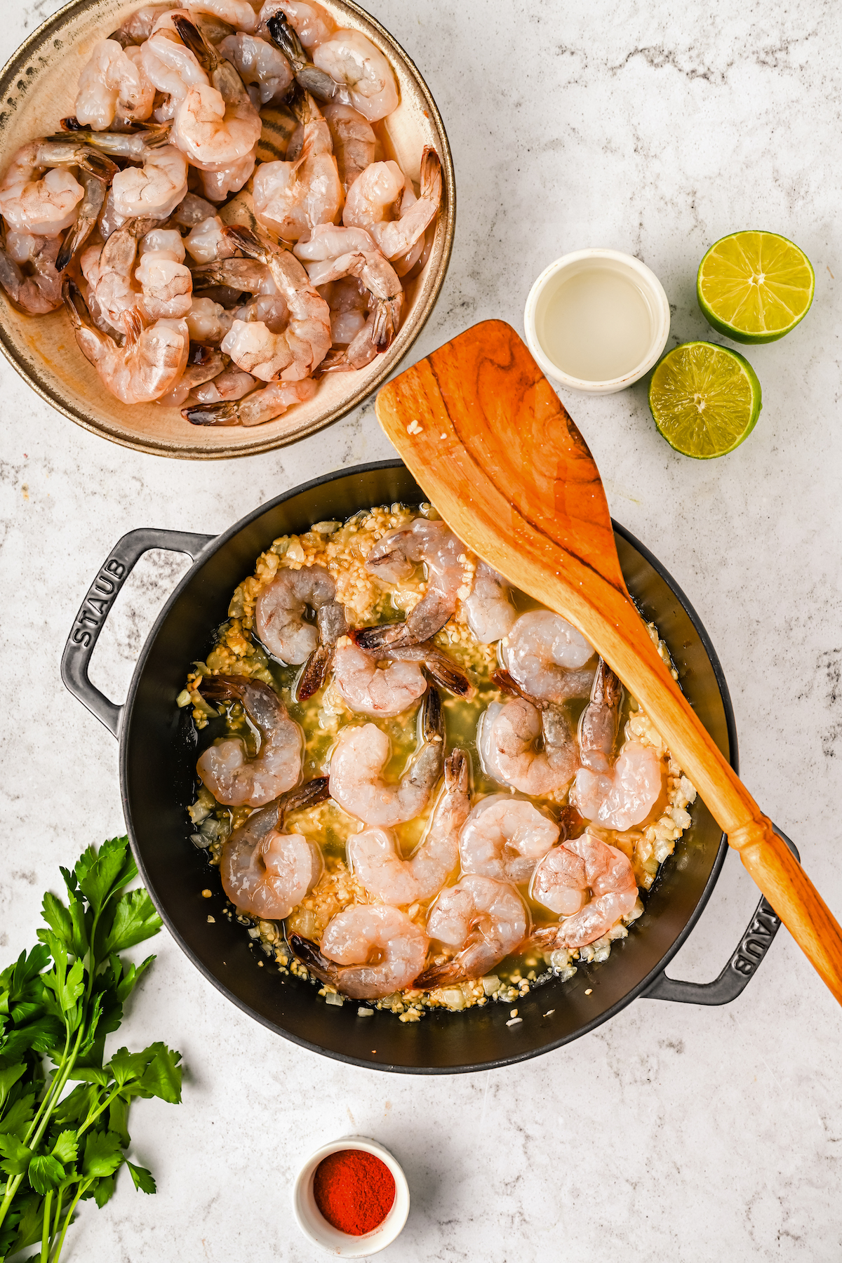 Raw shrimp cooking in a pot with garlic and onion and a wooden spoon, with a bowl of raw shrimp, a sprig of parsley, and a cut open lime on the table