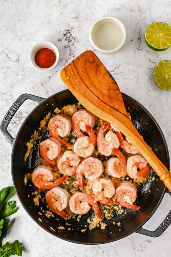 Shrimp cooking in a pot with onions and garlic, with a wooden spoon over the top, lime, vinegar, and paprika on the side, and a side of parsley