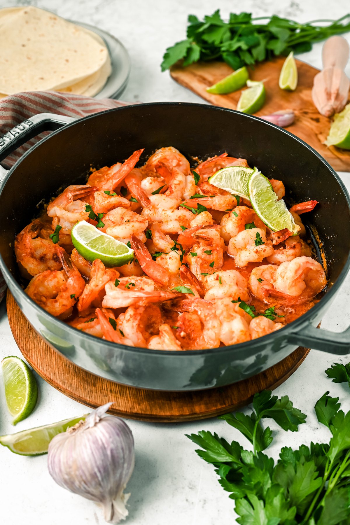 A pot of Camarones al Mojo de Ajo with lime slices on top, surrounded by lime slices, garlic bulbs, parsley, and fresh tortillas