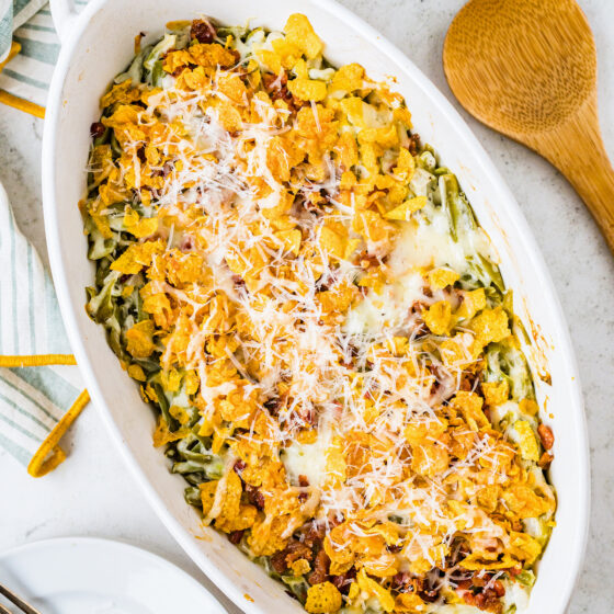 An oval casserole dish with a green bean casserole in it, topped with cornflakes and Parmesan.
